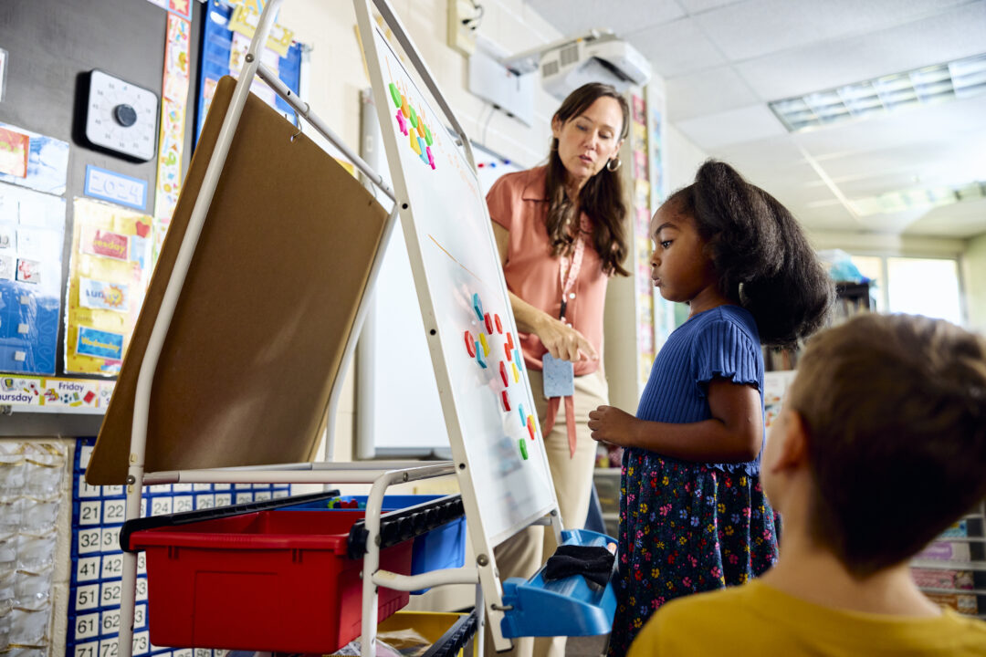 Teacher in the background and learner in the foreground look on as a learner stands in front of a magnetic easel to arrange colored shapes as part of a learning exercise