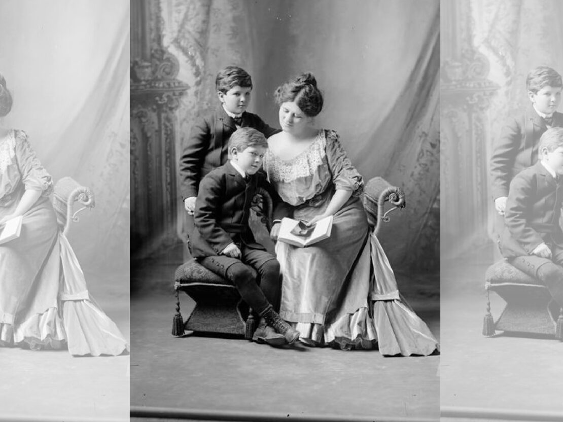 A black and white portrait of Belle wearing a formal dress, seated, with her sons next to her, looking at a book she is holding in her lap.