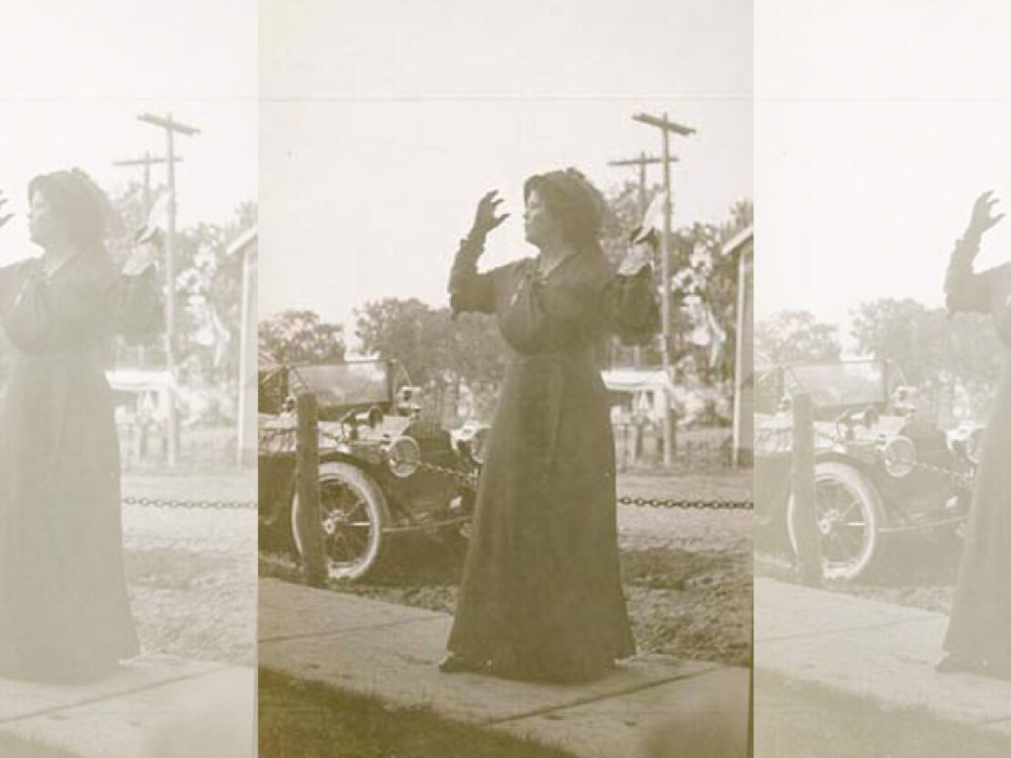 This black and white photo shows Belle Case La Follette dressed in a long dress with a hat, standing on a sidewalk with her arms raised, looking to the viewer's left.