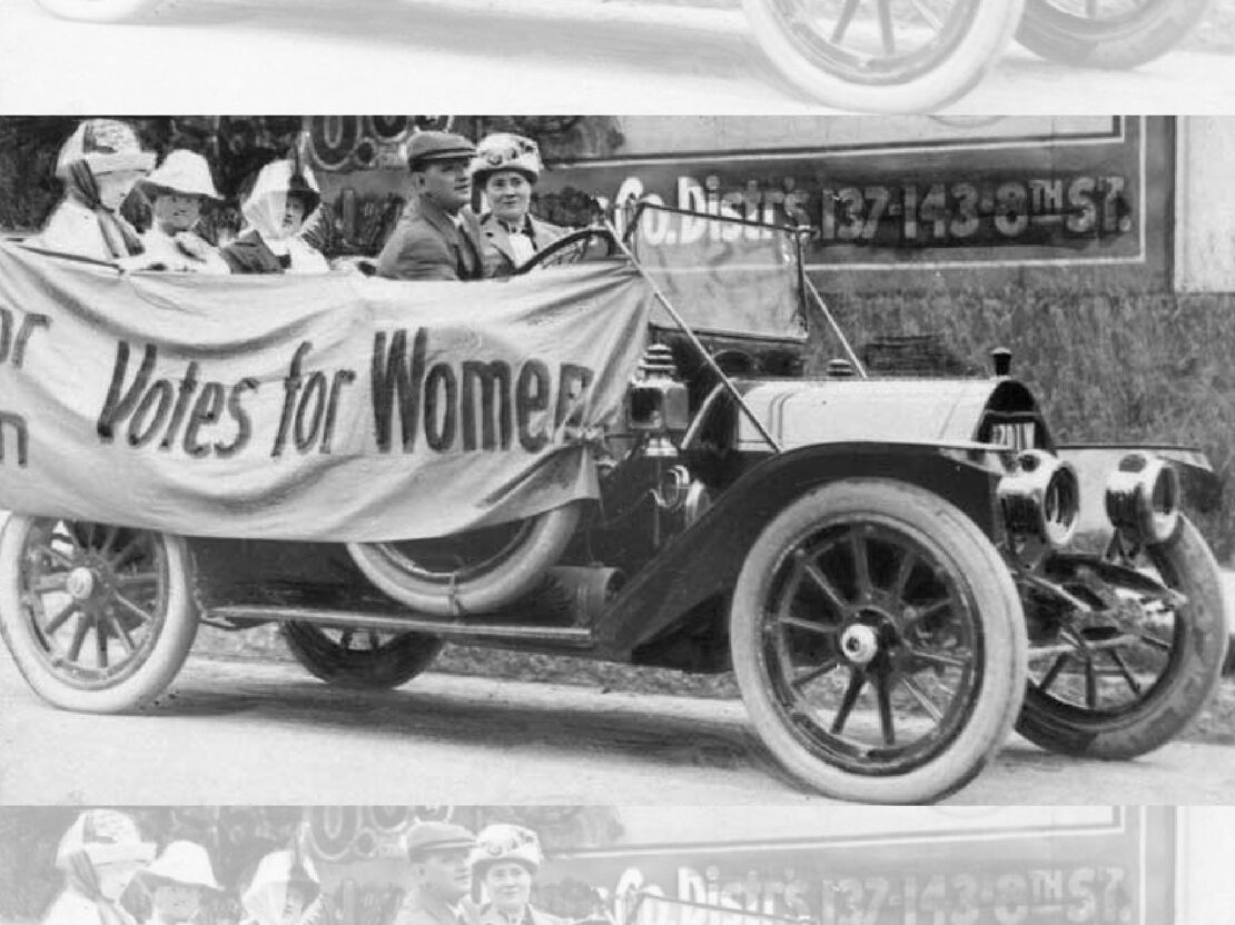 A black and white photo of a car with a banner reading "Votes for Women" hanging across the side of it. A man is sitting behind the wheel of the car. A woman is seated beside him and three women sit in the back seat.
