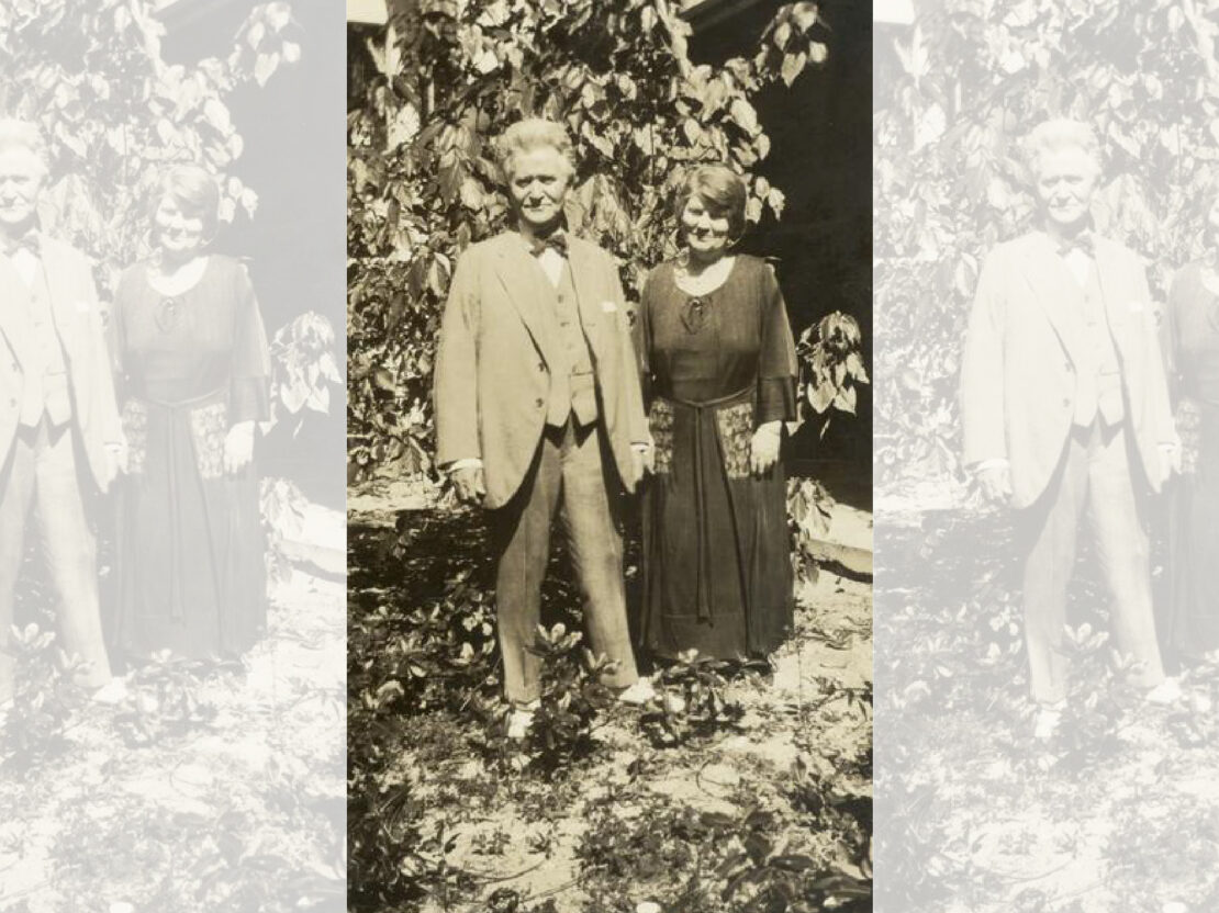 A black and white photo of Bob and Belle as older adults standing next to each other, holding hands, in front of a tree. Both have slight smiles on their faces. Bob wears a suit and Belle wears a looser-fitting long dress.