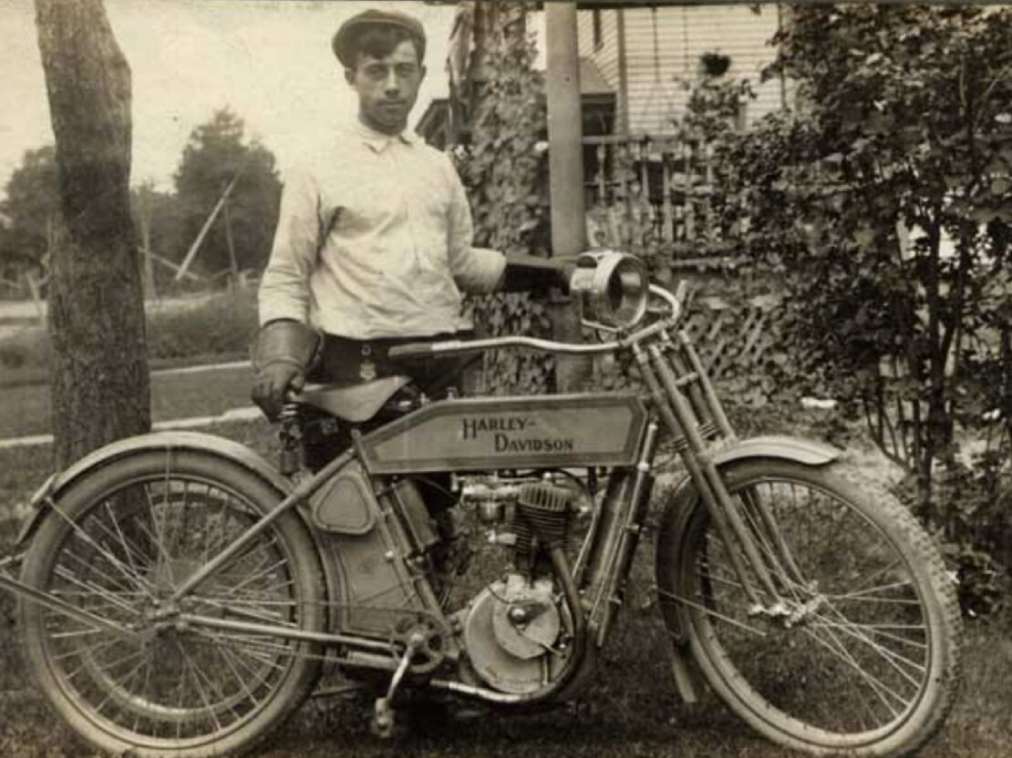 A black and white photograph of a man standing behind a motorcycle. On the side of the motorcycle are the words "Harley-Davidson."