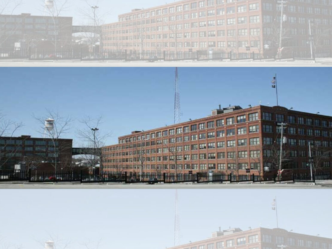 A color image showing a large red brick building with windows and a bright blue sky above it.