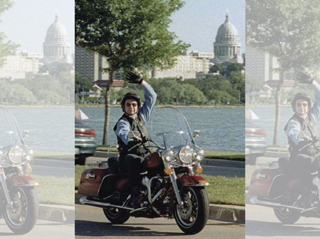 A color photograph showing a man riding a motorcycle on the road, with water and the state capitol in the background.