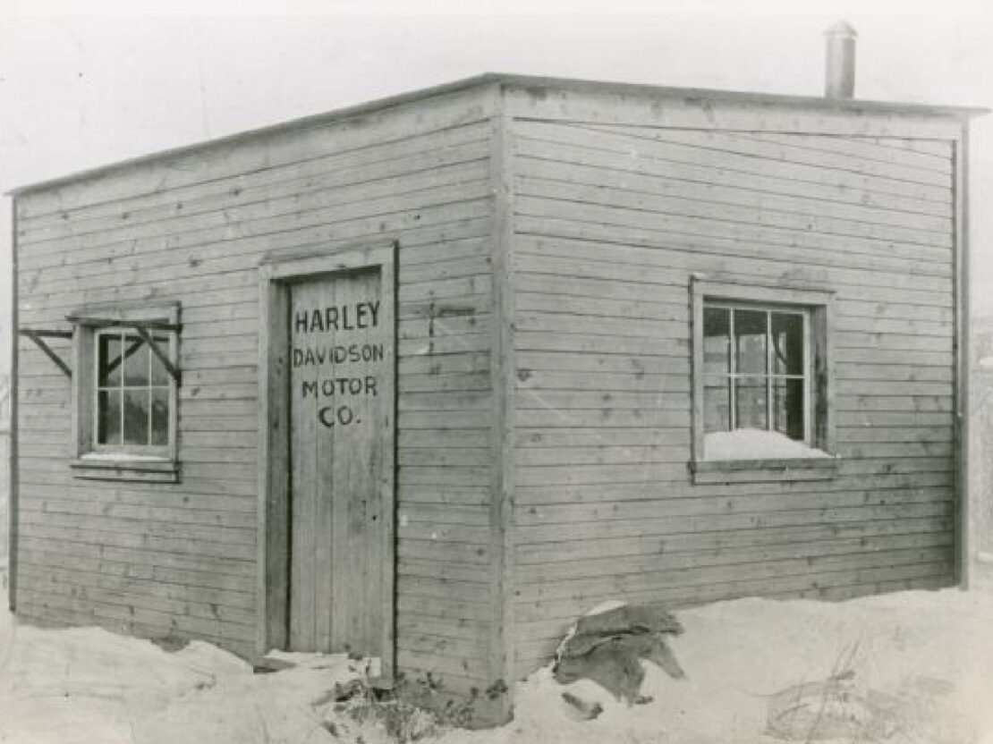A black and white photograph taken during the winter, showing the wooden backyard shed where the first Harley-Davidson motorcycles were assembled.