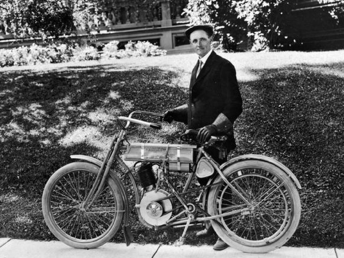 A black and white outdoor photograph of Walter Davidson standing on the edge of a sidewalk behind a motorcycle.