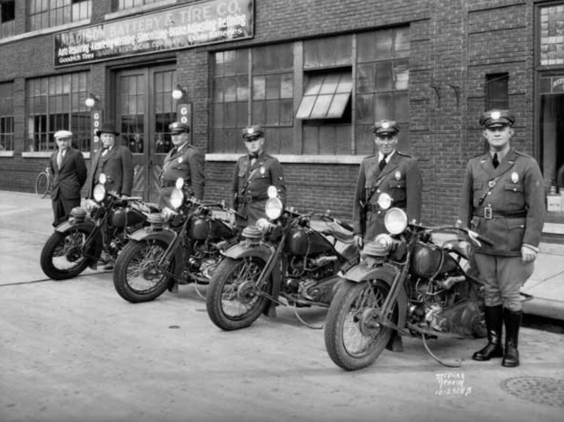 A black and white photograph showing a row of police officers, each standing next to a motorcycle.