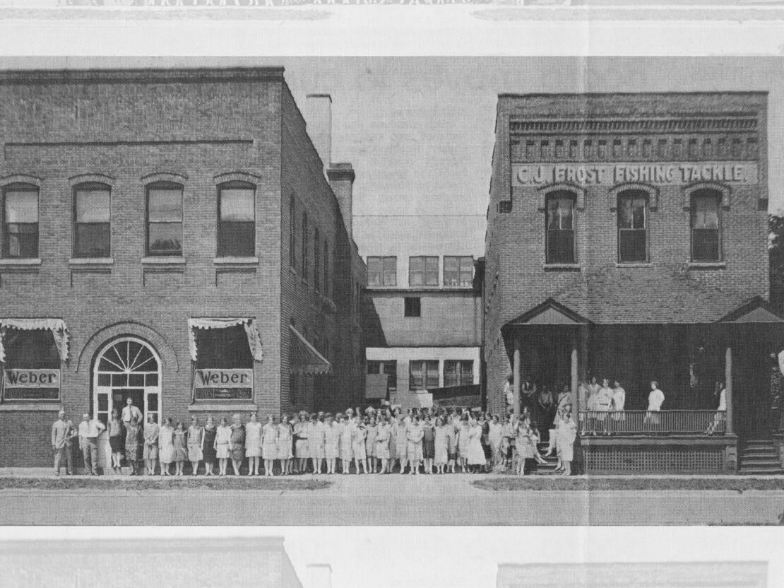 A black and white photo of two connected brick buildings, one with the name C.J. Frost Fishing Tackle on it, and the other with “Weber” appearing in the windows. A group of women are standing in front of the buildings.
