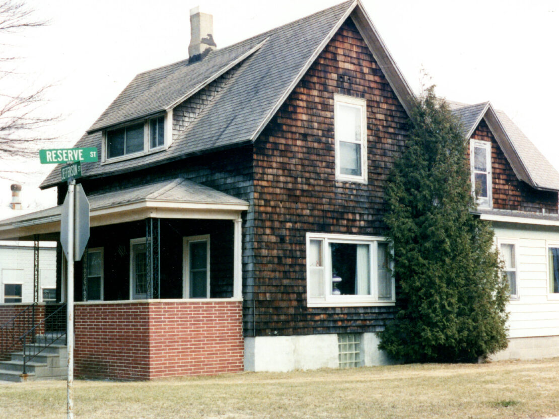 A color photo showing a house with brown shake siding, with the street sign showing “Reserve St” and “Jefferson St” in the left side of the photo.