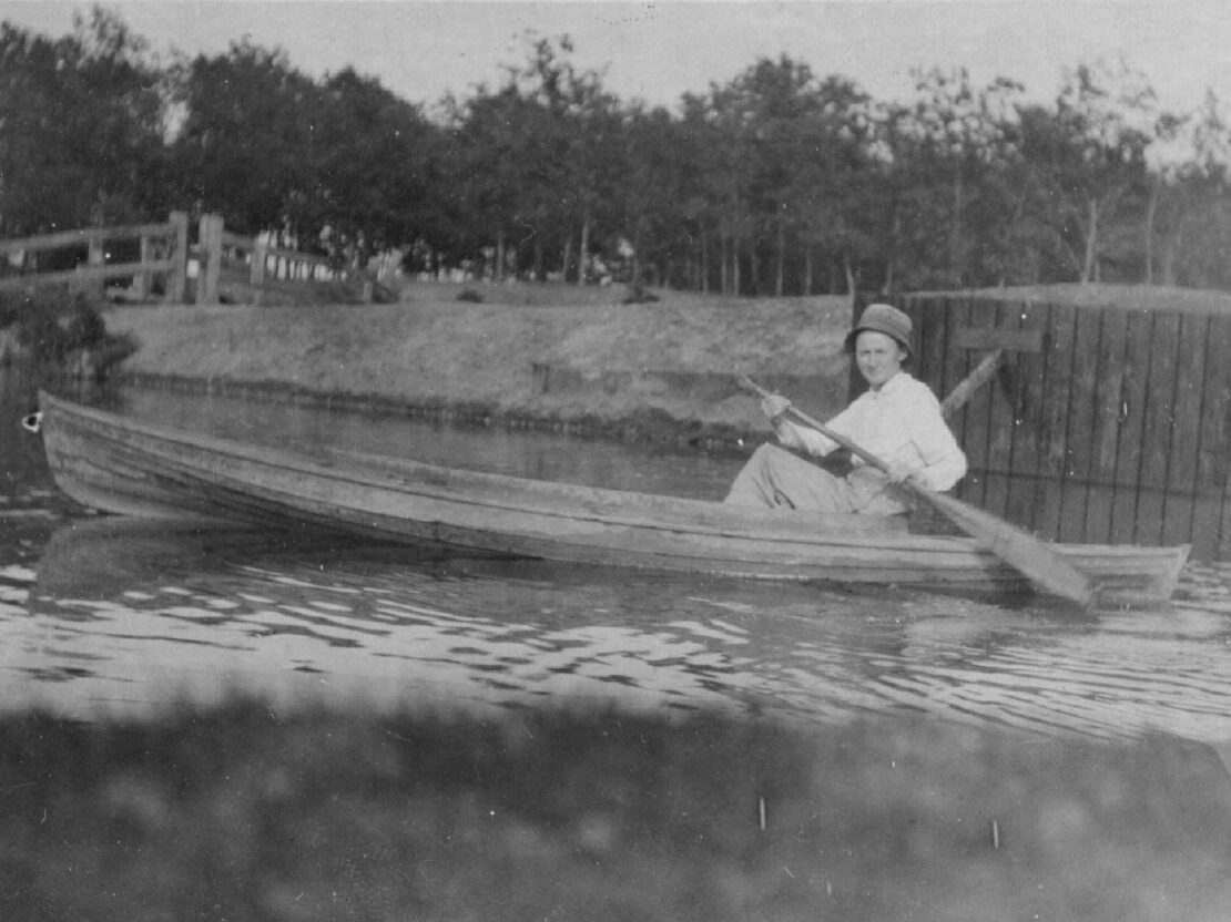 A black and white photo of Carrie sitting in a canoe on the water, holding a paddle in her hands and looking towards the viewer.