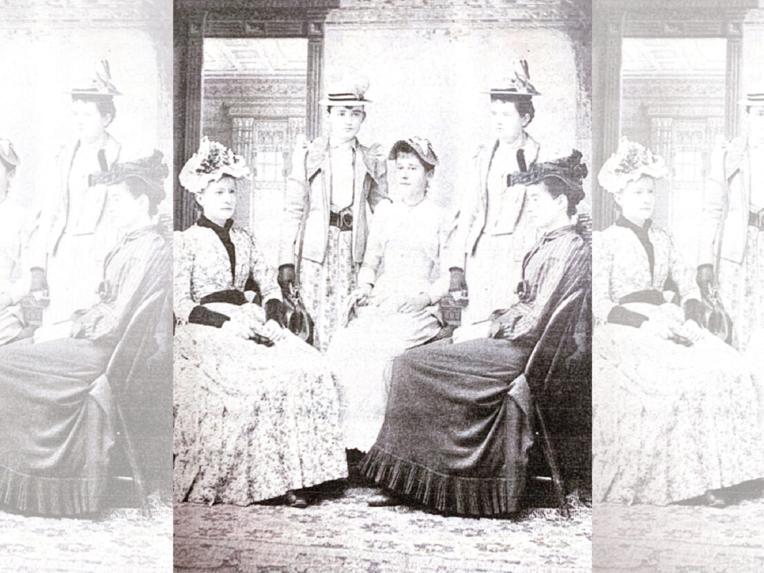 A black and white photograph of Carrie posing with her sister and three friends in a formal room inside a building. All are wearing hats and long skirts, and looking in different directions.