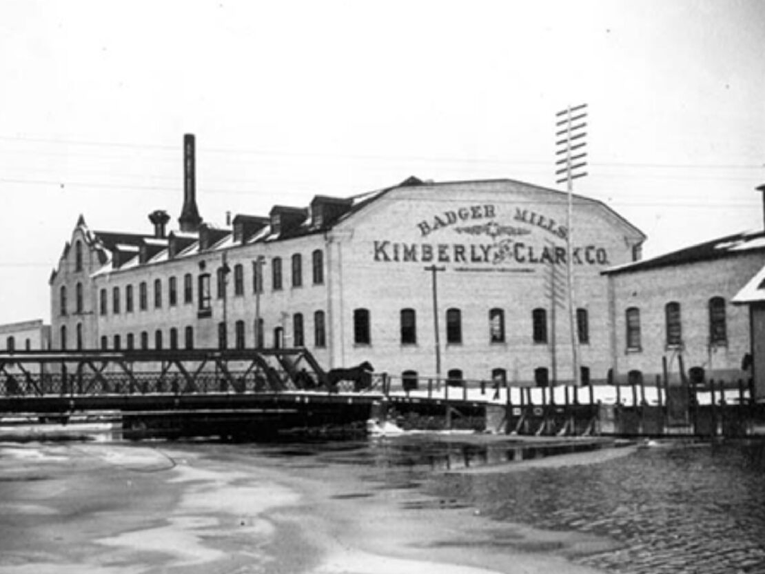 A black and white photograph showing a large mill building behind a bridge and a body of water.