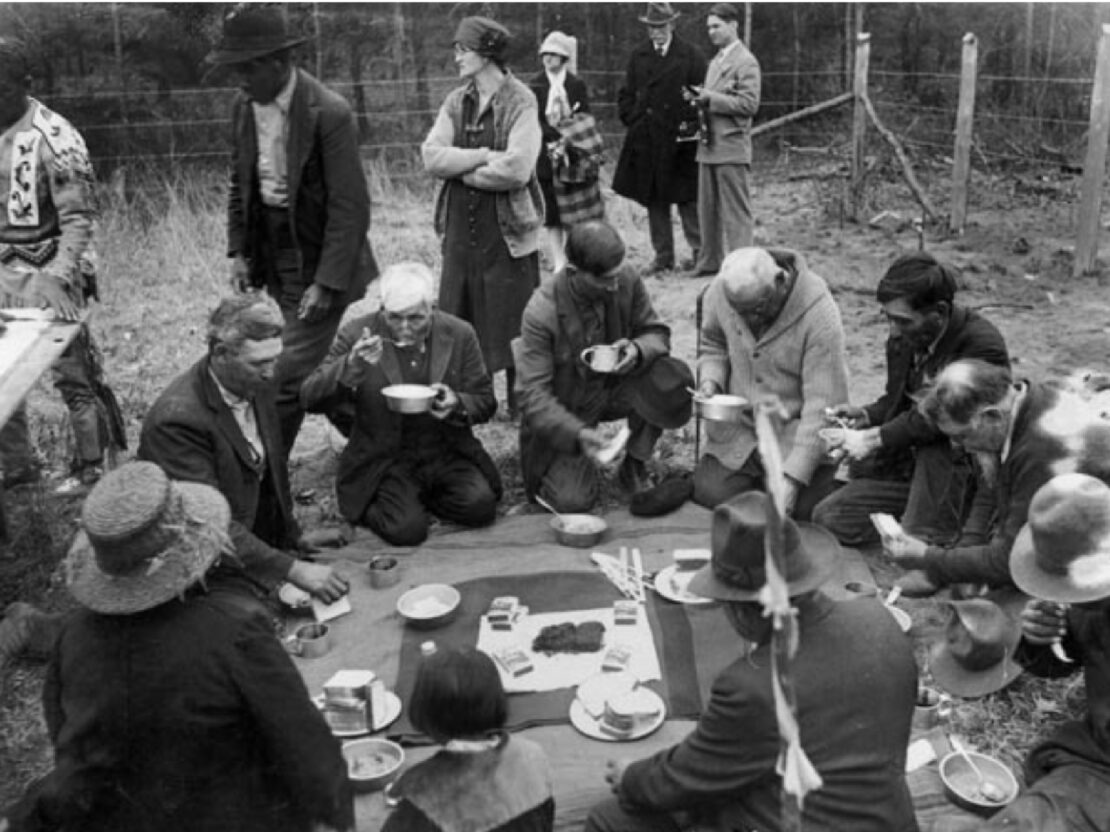 A black and white photo of people kneeling next to each other, eating from bowls.