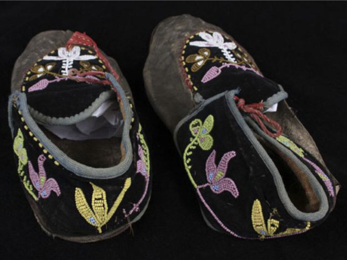 A color image showing a pair of small black moccasins with purple, green, white, read and yellow beading on them.