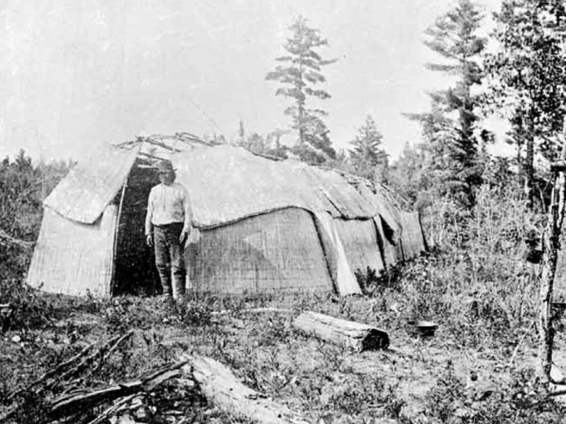A black and white photograph of a person standing outside of a structure with a rounded roof, next to a structure made of two beams, with grass and trees behind them.