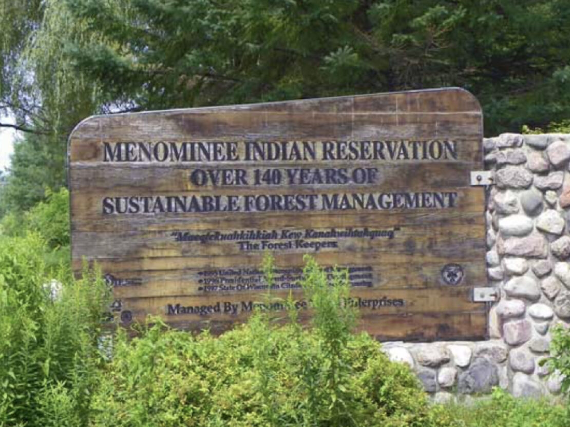 A color image of a wooden sign with text on it. The heading reads, “Menominee Indian Reservation Over 140 Years of Sustainable Forestry Management.”
