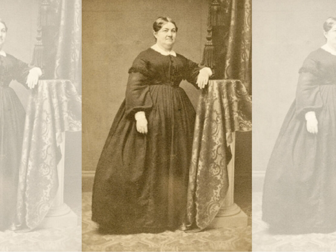 This black and white photograph is a formal portrait of Elizabeth Baird when she was an adult. She poses in a long dress, with her left arm resting on a covered stand, and looks to the right of the viewer.