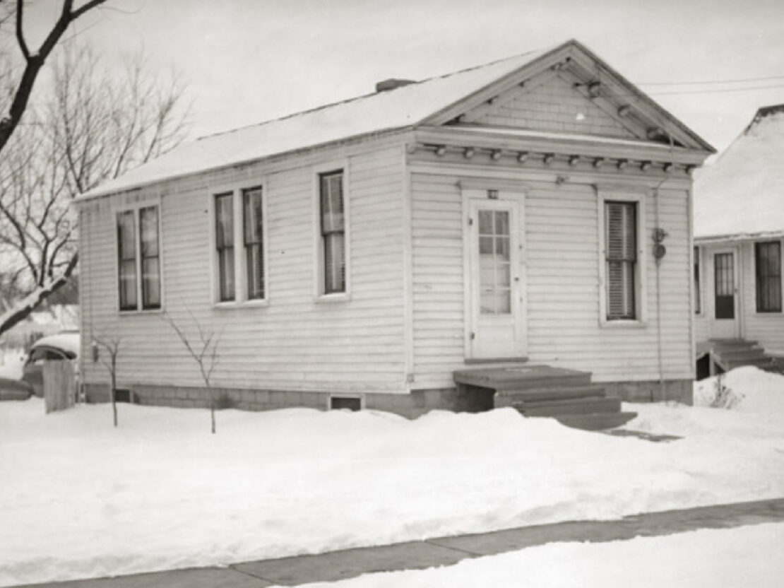 A black and white photograph of a small white house sitting in a snow-covered lawn, with a cleared sidewalk, tree, and small white house near it.