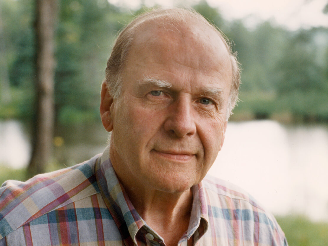 A full color photograph of Gaylord Nelson as an older adult. His head and shoulders are visible and he is looking directly at the viewer. He wears a multicolor plaid shirt. Behind him are trees and water.