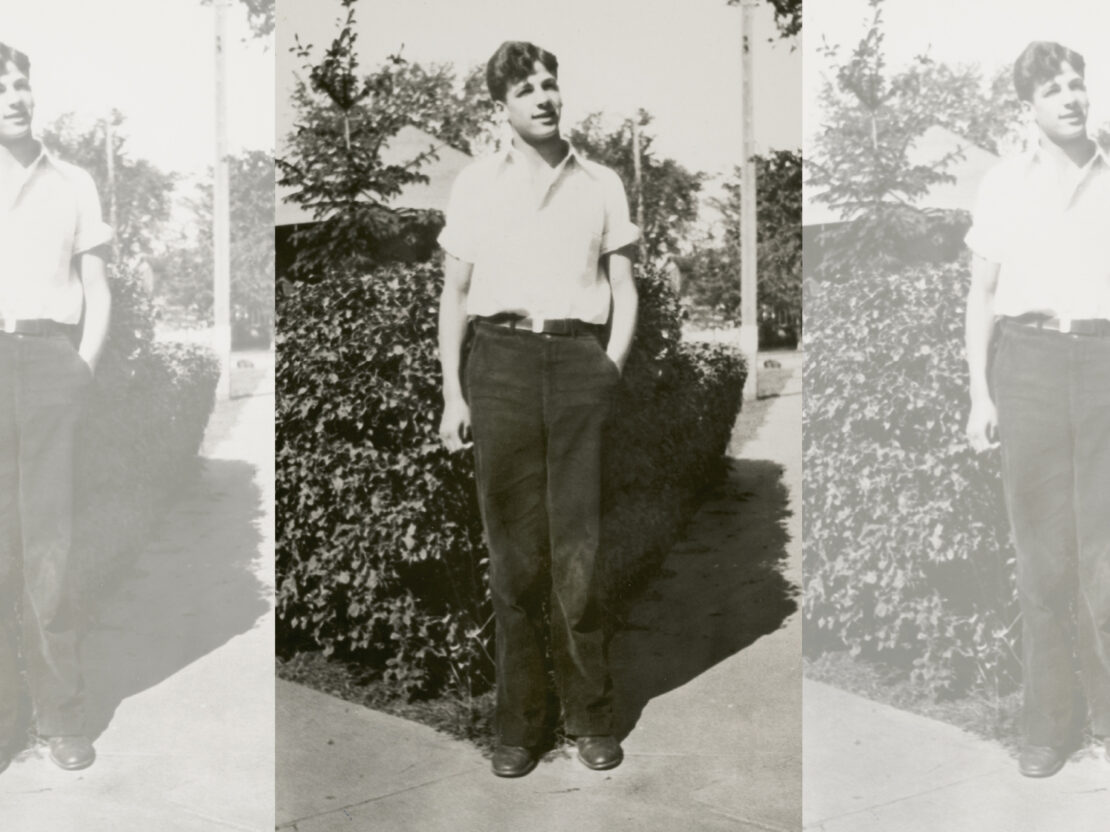A black and white photograph of Gaylord Nelson. He stands on the sidewalk in front of a shrub. He is wearing a plain collared shirt and pants. He is looking to the right of the viewer and has his left hand in his pocket and his right arm at his side.