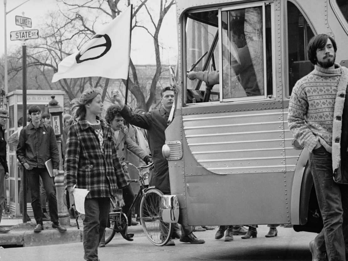 In this black and white photograph, several young people are walking together. One person is carrying a flag with the ecology symbol on it. Part of a bus is also visible.