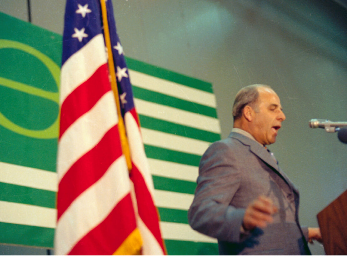 A color image showing Gaylord Nelson speaking at a podium, with a United States flag and ecology flag behind him.