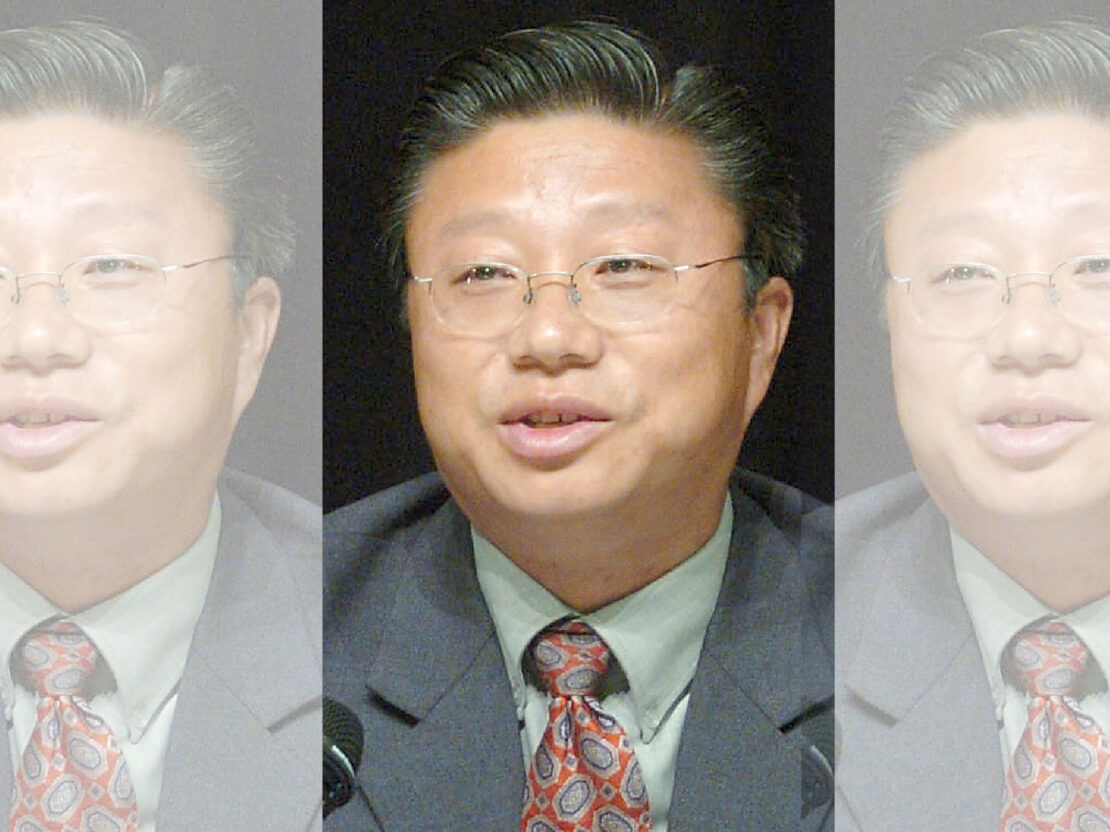 A color photograph of Joe Bee Xiong sitting in front of a microphone. He is wearing a suit and glasses. He is looking to the left of the viewer.