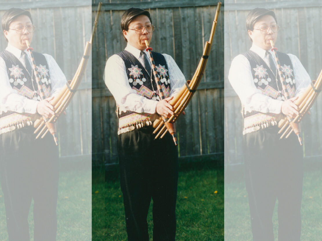 A color image showing Joe Bee standing and playing the qeej outside.