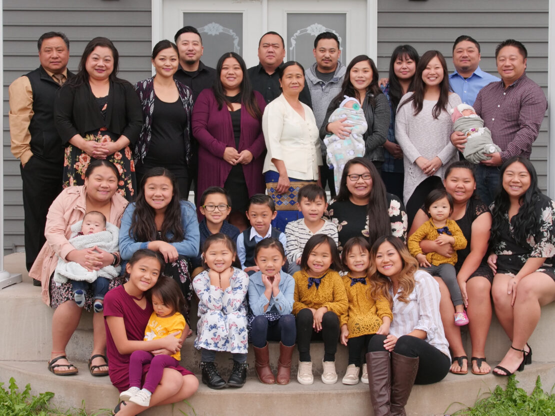 A color photograph of the Xiong Family. The top row of adults is standing, while the middle and bottom rows of children and young adults are sitting on the concrete steps of a porch to a building.