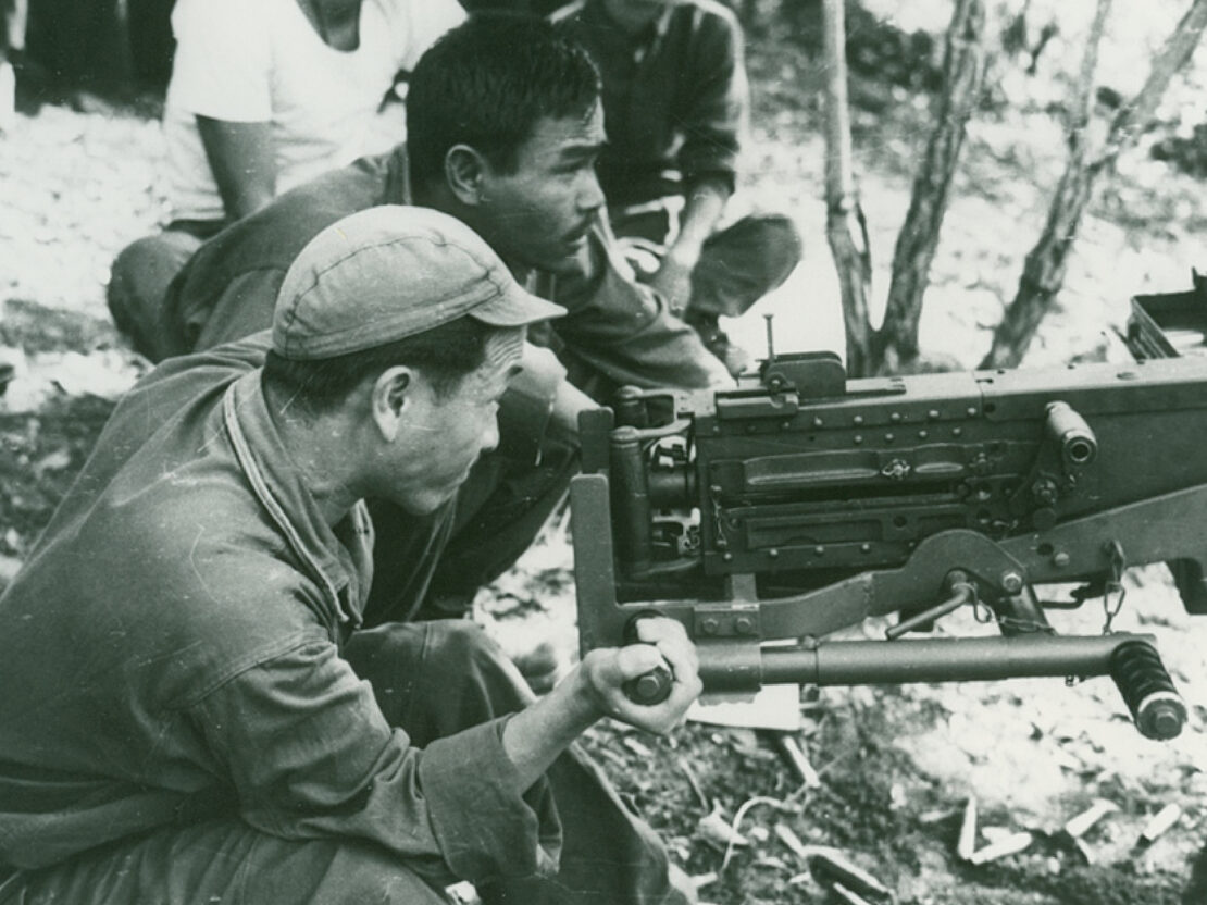 A black and white image showing two men crouched behind a large weapon.