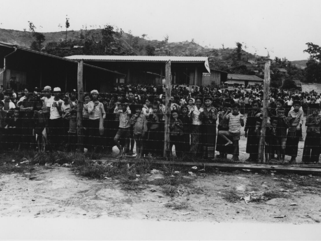 A black and white image of a crowd of people standing behind a wire fence.