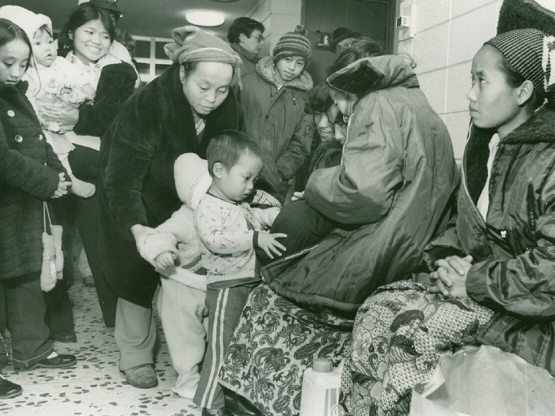 A black and white image showing two seated people and several standing people, all wearing coats.