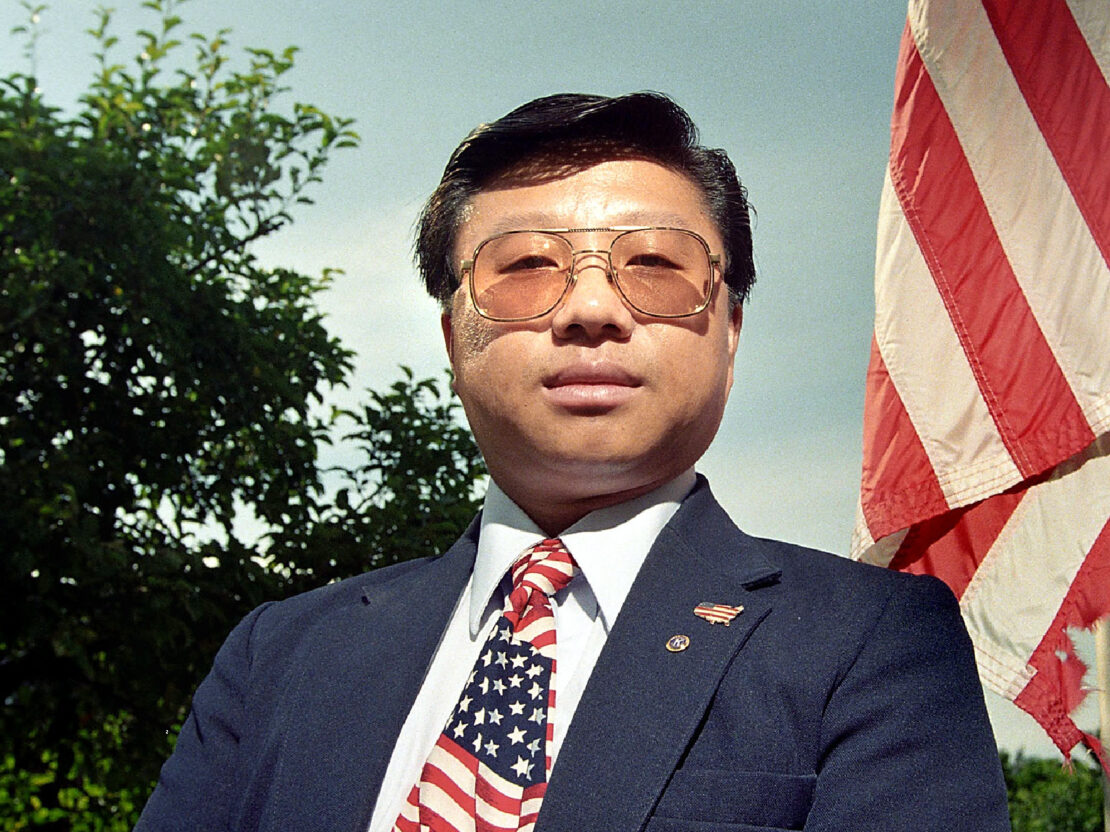 A color photograph of Joe Bee Xiong standing in front of an American flag, wearing a suit with an America flag-patterned tie. In the photo his head and shoulders are visible and he is looking at the viewer.