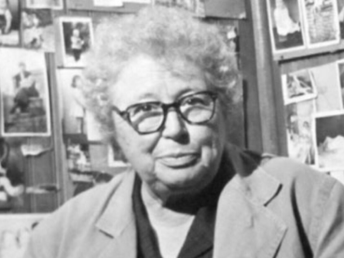 A black and white photograph of Dr. Kate Pelham Newcomb. She is looking at the viewer and behind her are lots of pictures on the walls.