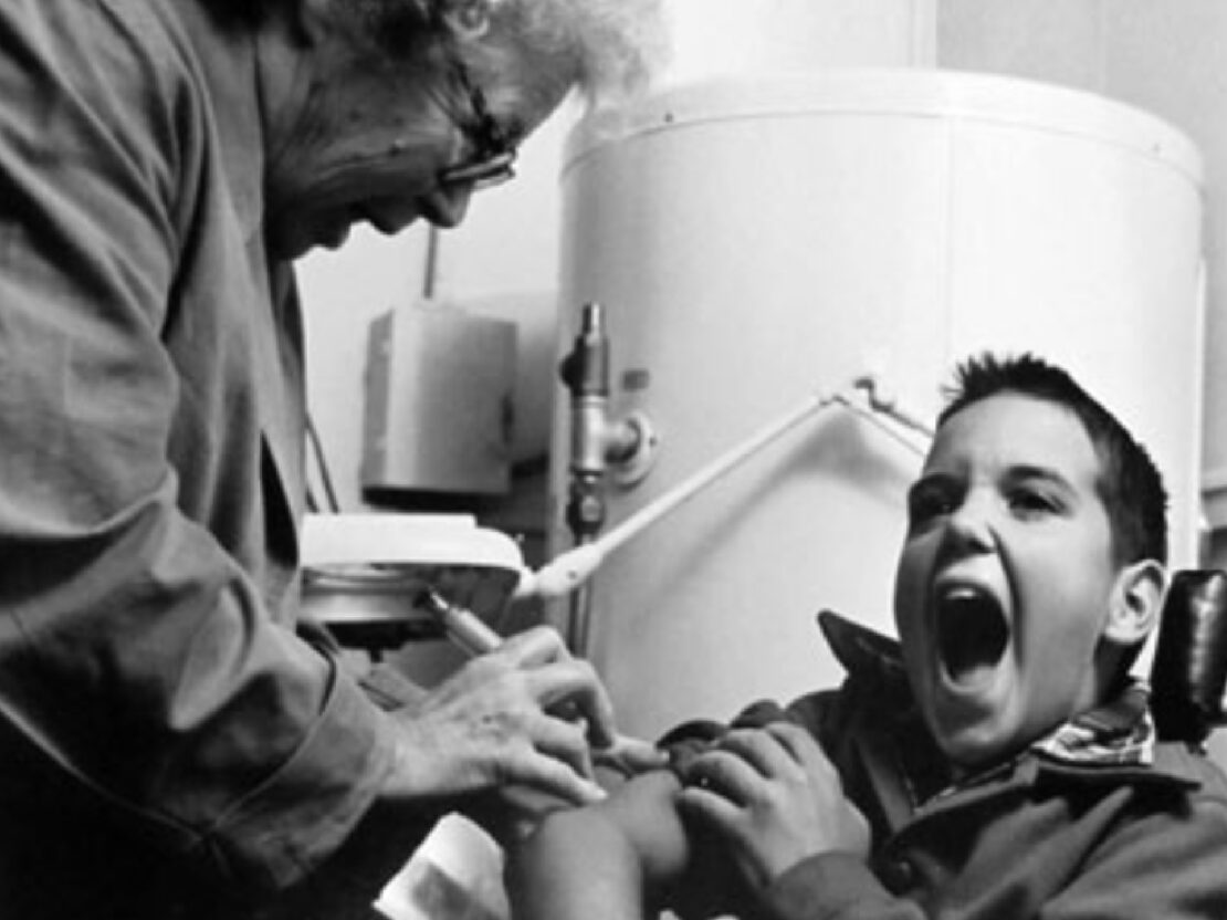 A black and white image of Doctor Kate giving a shot to a boy in his arm. He has his mouth open, in reaction to receiving the shot.