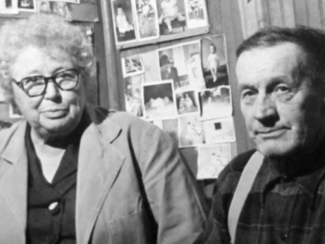 A black and white photograph showing Kate and Bill next to each other in front of a wall of photographs.