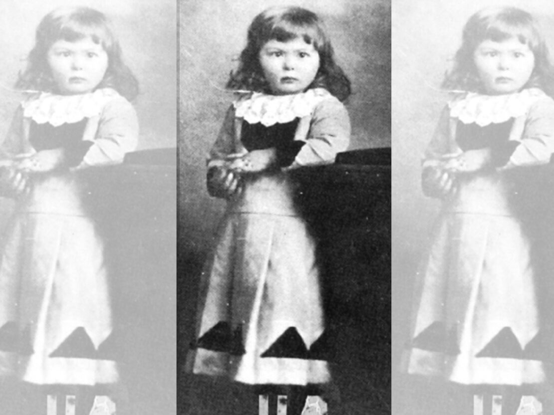 A black and white photograph of young Kate standing, with her left arm propped on a stand. She looks to the right of the viewer with a serious expression on her face.