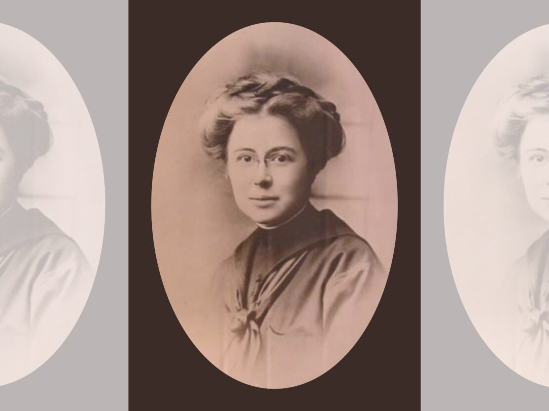 A black and white photograph with a reddish hue, showing Kate Pelham. In the photograph her head and shoulders are visible, and she is looking directly at the viewer.