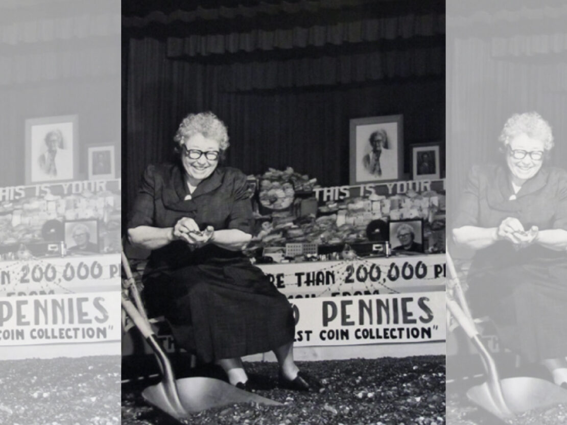 A black and white photograph showing Kate seated, holding something in her hand and smiling. Next to her is a table with memorabilia and a sign with the word “Pennies” visible. Surrounding her feet are lots of pennies.
