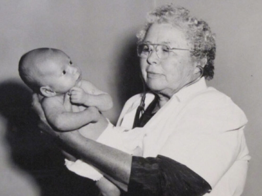 A black and white photograph of Dr. Kate standing with her arms outstretched, cradling a baby.