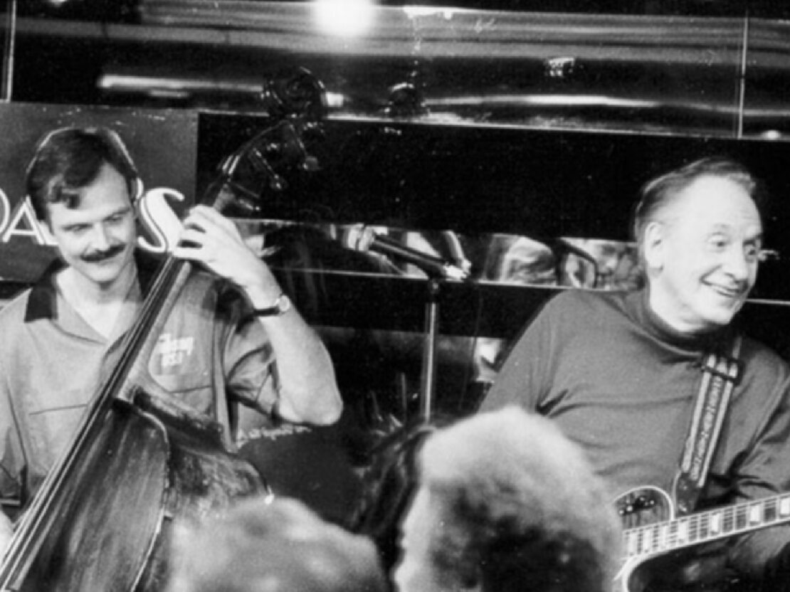 A black and white photograph of a much older Les on stage with a band playing music. Les is smiling.