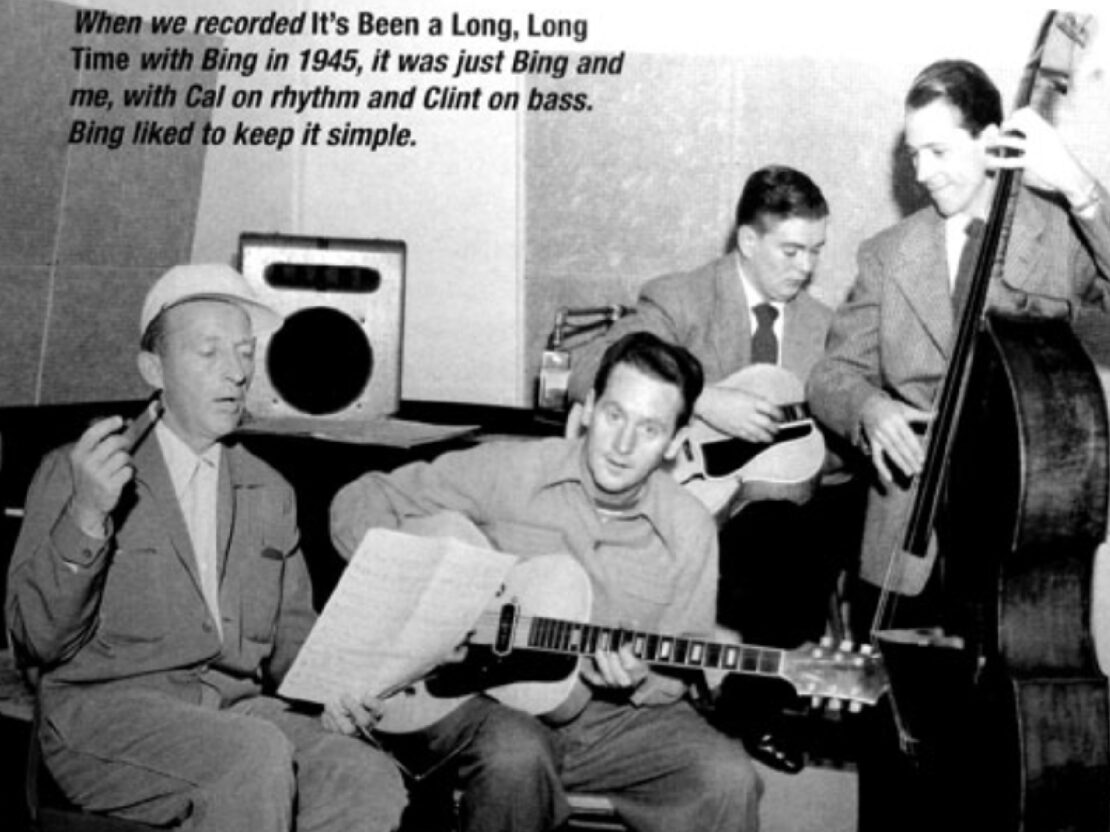 A black and white photograph in the studio. Les is seated in the center of the photo, holding a guitar and looking at the viewer. On the left side of the photo is Bing Crosby, who is looking down at a piece of paper. On the right side of the photo are Les's bandmates with a guitar and a string bass.