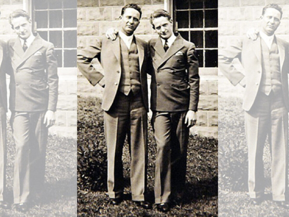 A black and white photograph of two men standing next to each other, wearing suits, looking at the viewer.