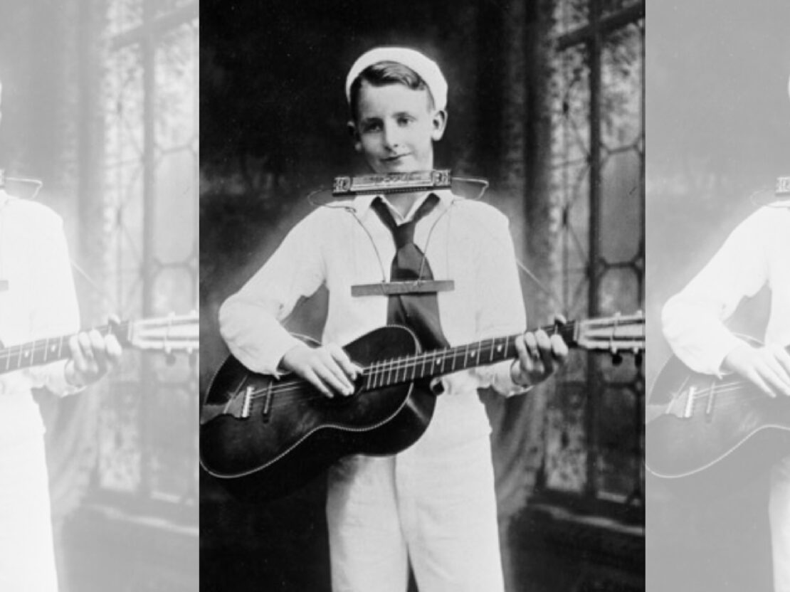 A black and white photograph of young Les Paul standing and playing the guitar and harmonica, with a wire holding the harmonica in place.