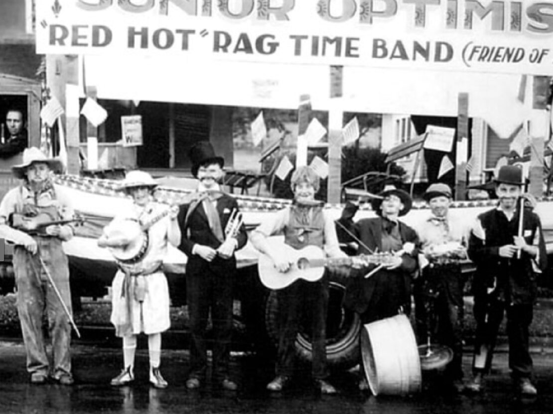A black and white image of a group of young people with instruments standing in front of a loat wtih a sign that reads, “Junior Optimist 'Red Hot' Rag Time Band.”