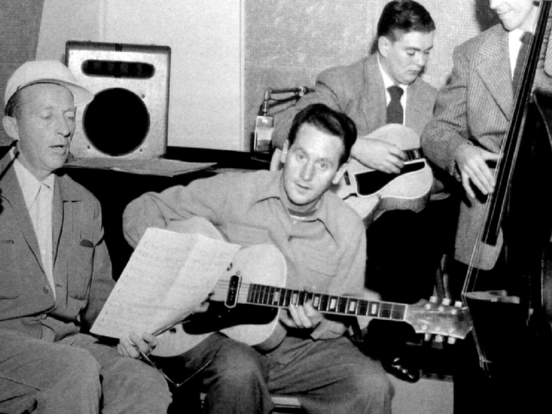 A black and white photograph in the studio. Les is seated in the center of the photo, holding a guitar and looking at the viewer. On the left side of the photo is Bing Crosby, who is looking down at a piece of paper. On the right side of the photo are Les's bandmates with a guitar and a string bass.