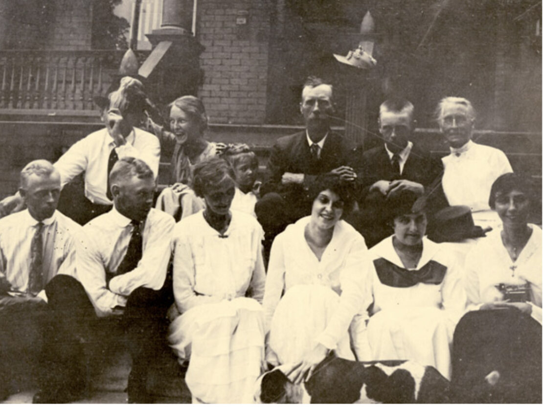 A black and white photograph of a group of people sitting together on the steps of a brick building.