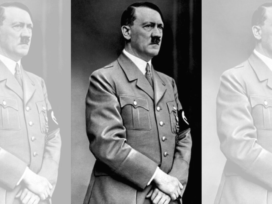 A black and white photograph of Adolf Hitler. He is standing in uniform and is looking to the right of the viewer.