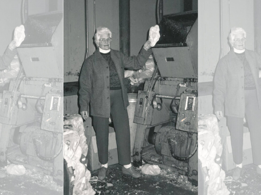A black and white image of Milly standing inside a warehouse holding up an item to go into the chute of the grinder.