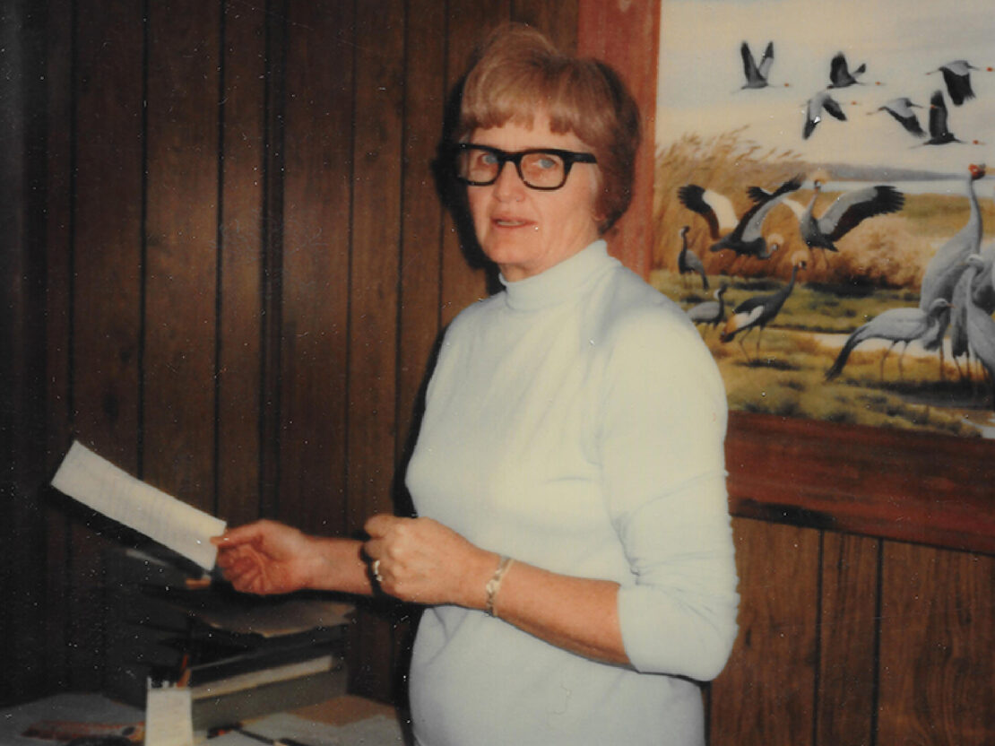 A color photograph of Milly standing in front of a wood paneled wall that has a framed picture of cranes on it. Milly is holding up a piece of paper and is looking towards the viewer.
